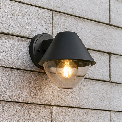 Schwarze Wandleuchte mit klarem Glasschirm und E27-Fassung, frontal fotografiert vor einer Hausfassade 